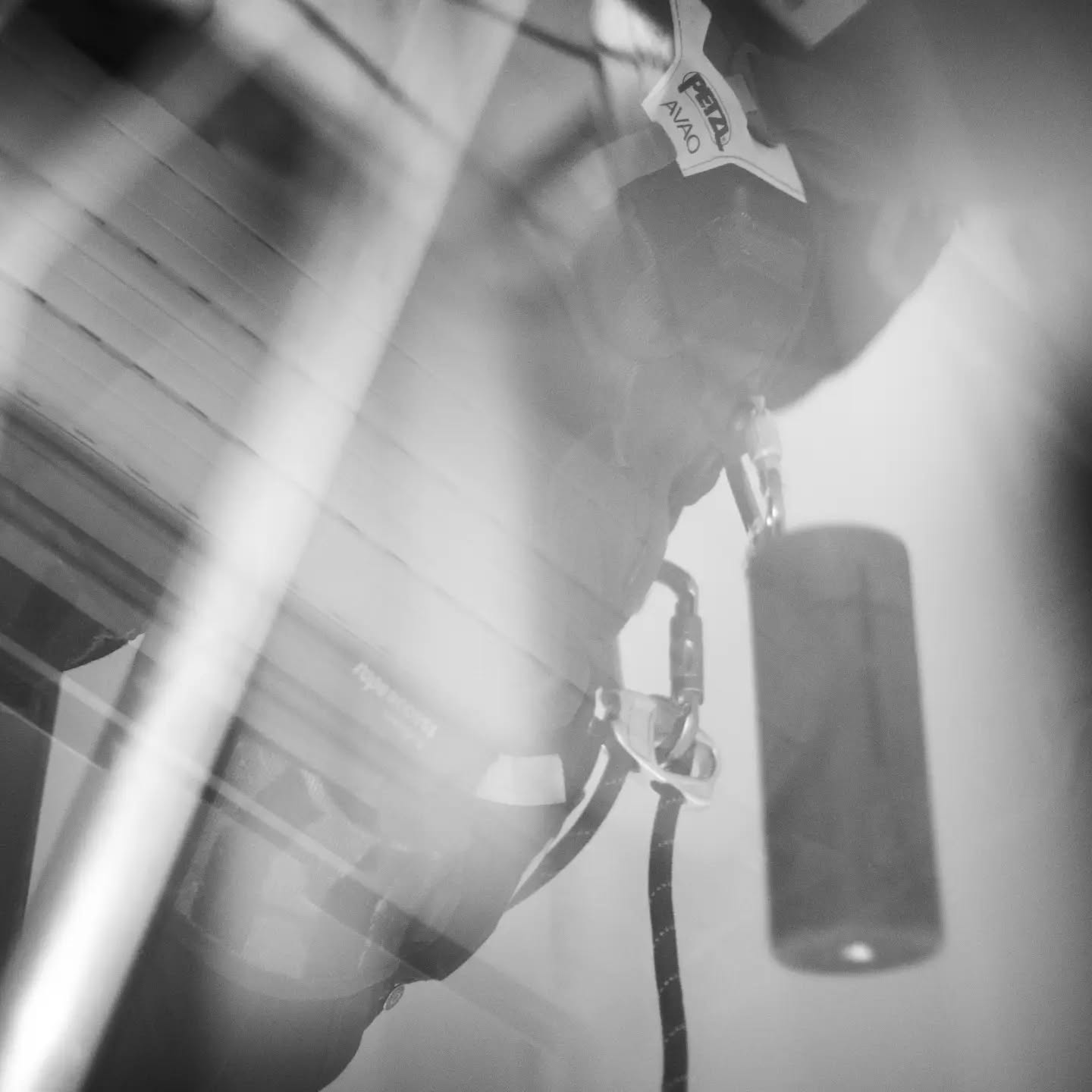 Black and white double-exposure image of a climbing harness with carabiners and Petzl Avao label, overlaid with a striped pat