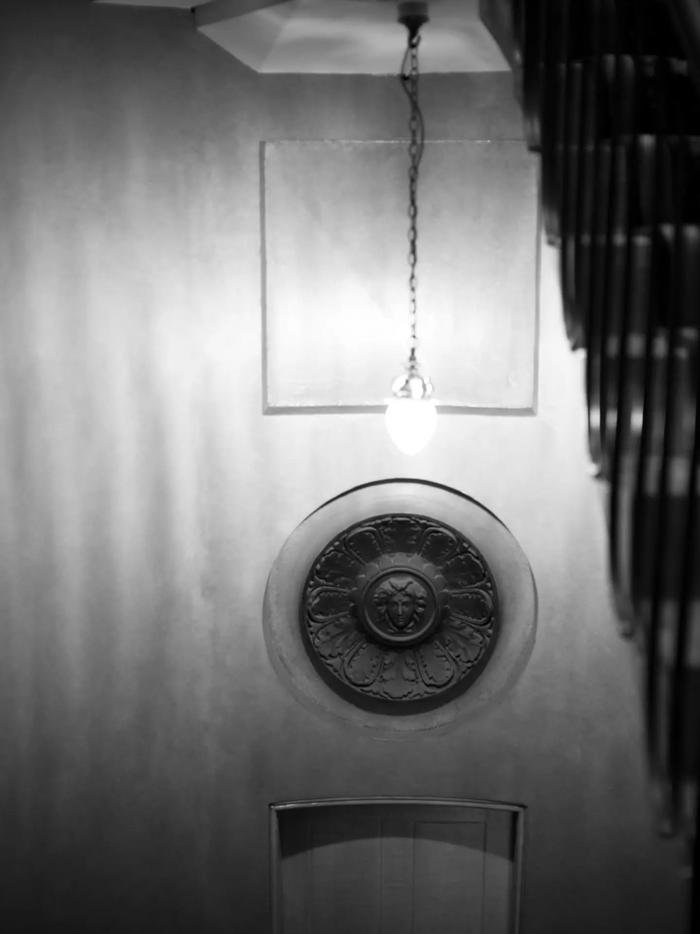 Black-and-white photo of a hallway with a hanging pendant light, a circular medallion with a face motif, and dark vertical ra