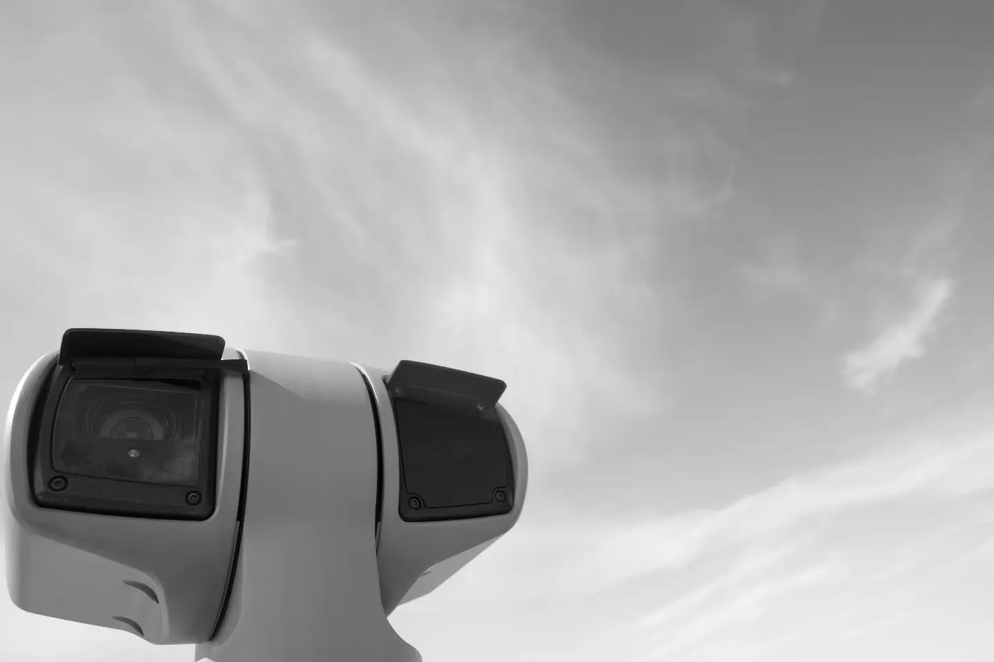 Black-and-white photo of a security camera housing with two lenses, shot from below against a cloudy sky.