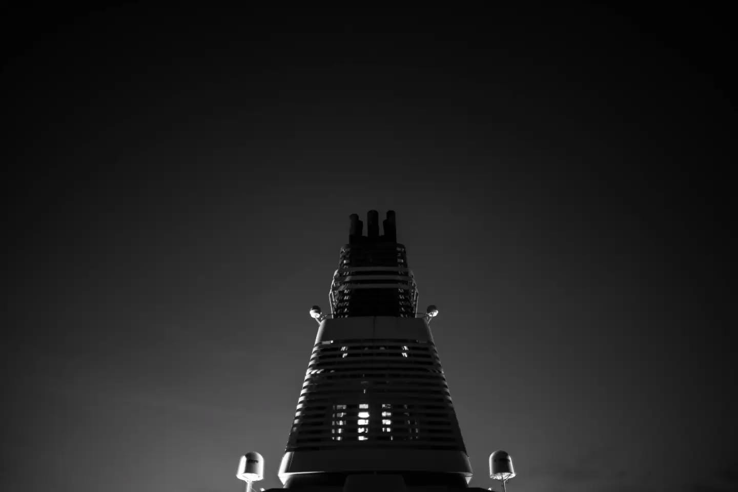 Black and white low-angle photo of a tapered tower with a ribbed exterior and multiple spires at its peak, against a dark sky
