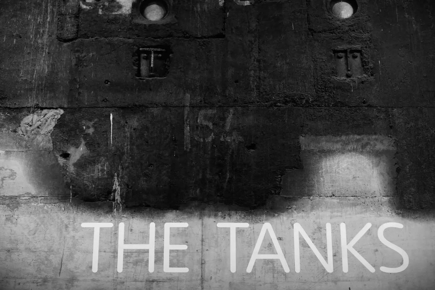 Black-and-white photo of a weathered concrete wall with round and rectangular metal fittings, streaked stains, and "THE TANKS" in white text below.