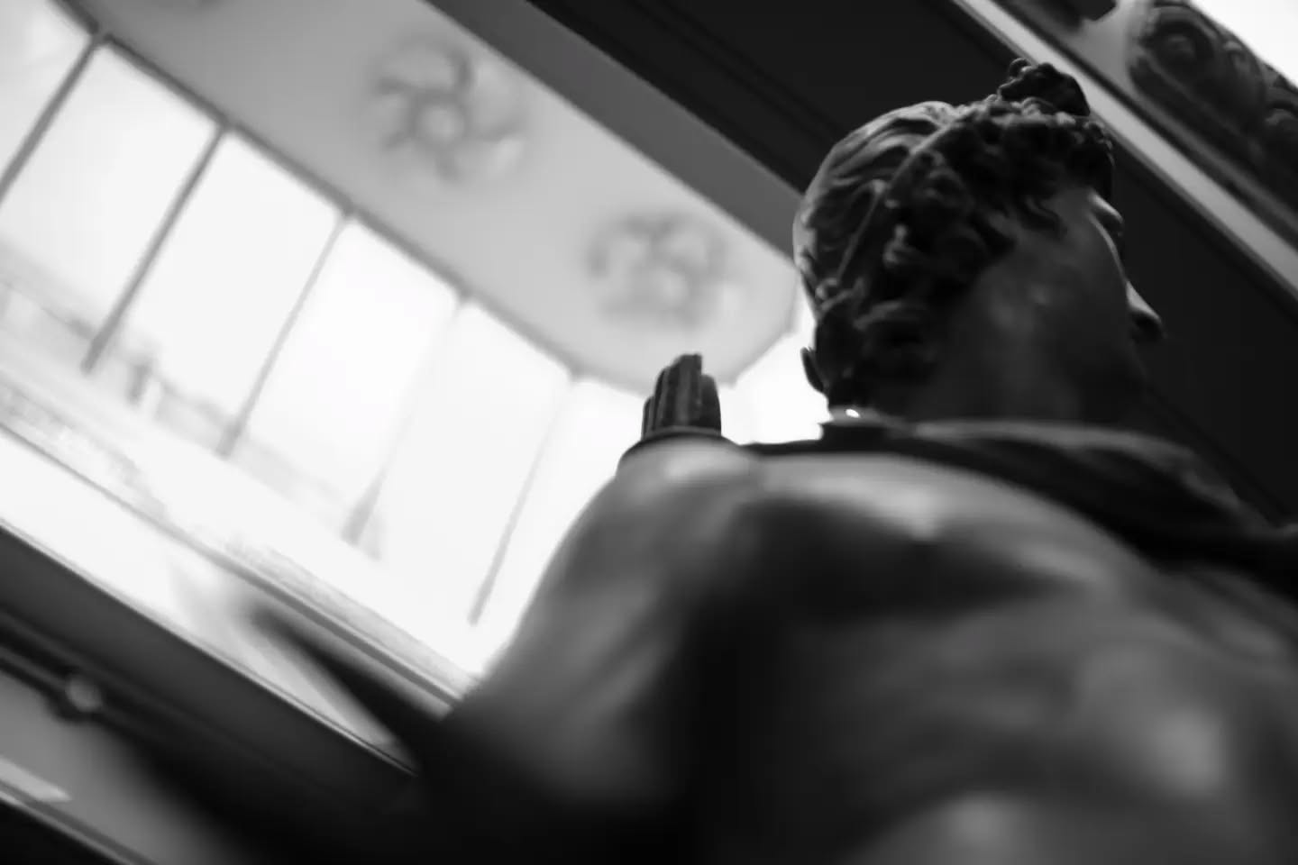 Black-and-white photo of a classical bronze statue's head and shoulders from behind, raised hand, bright window in background