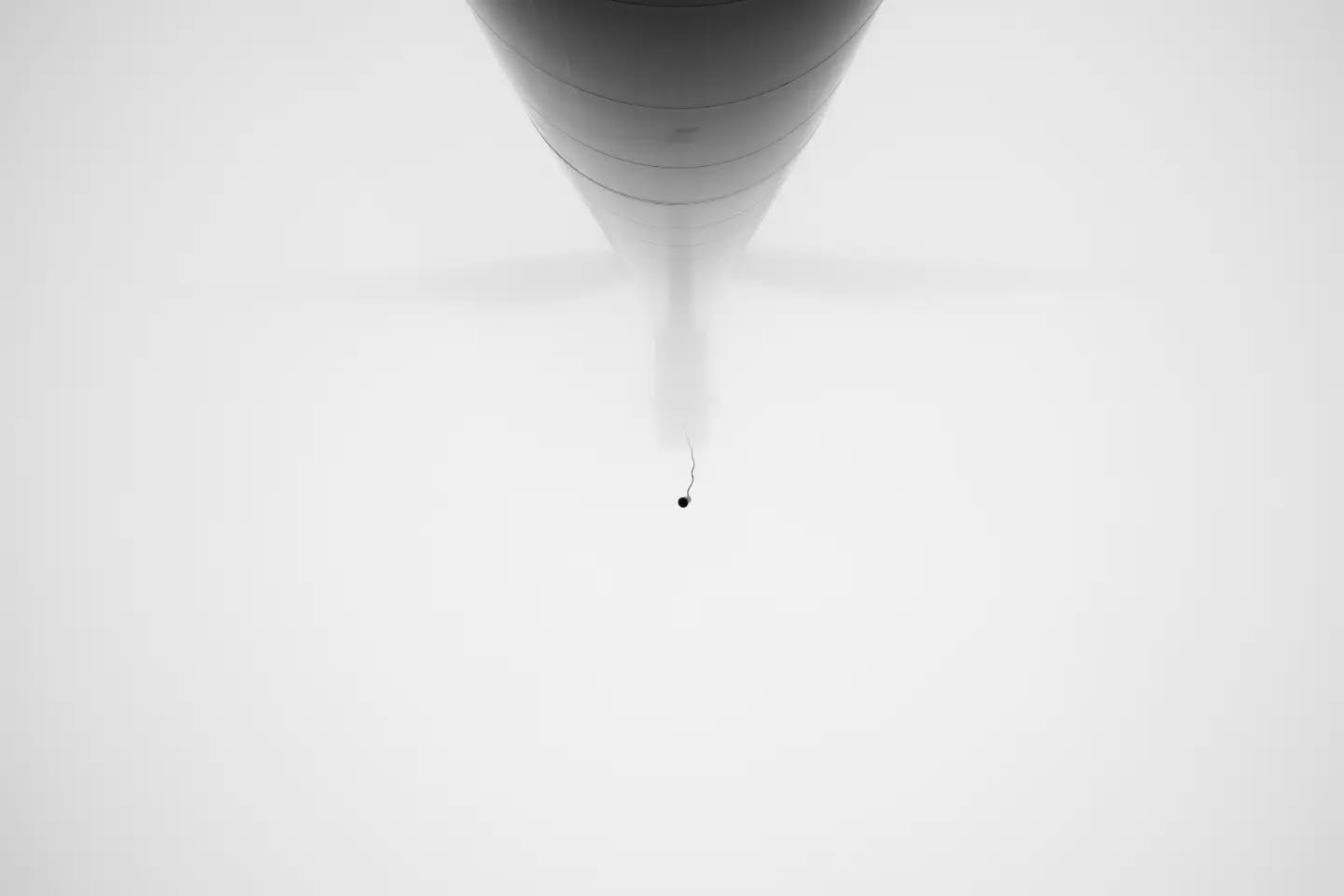 Black-and-white image of a hot air balloon's underside fading into fog, with a small dark weight hanging on a thin line below
