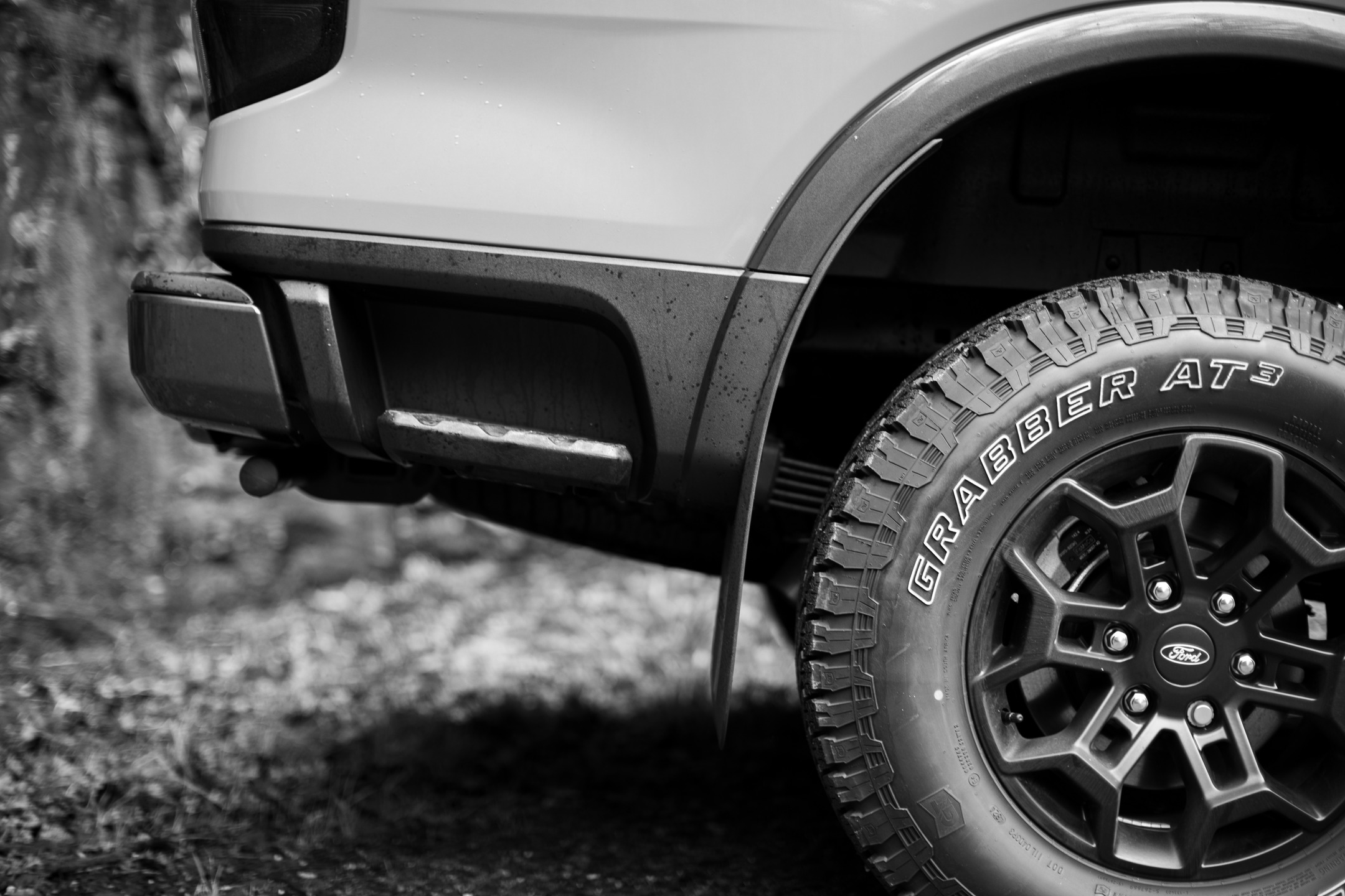 Black and white close-up of a Ford truck's front wheel, showing Grabber AT3 tire tread, alloy rim, and lower fender panel.