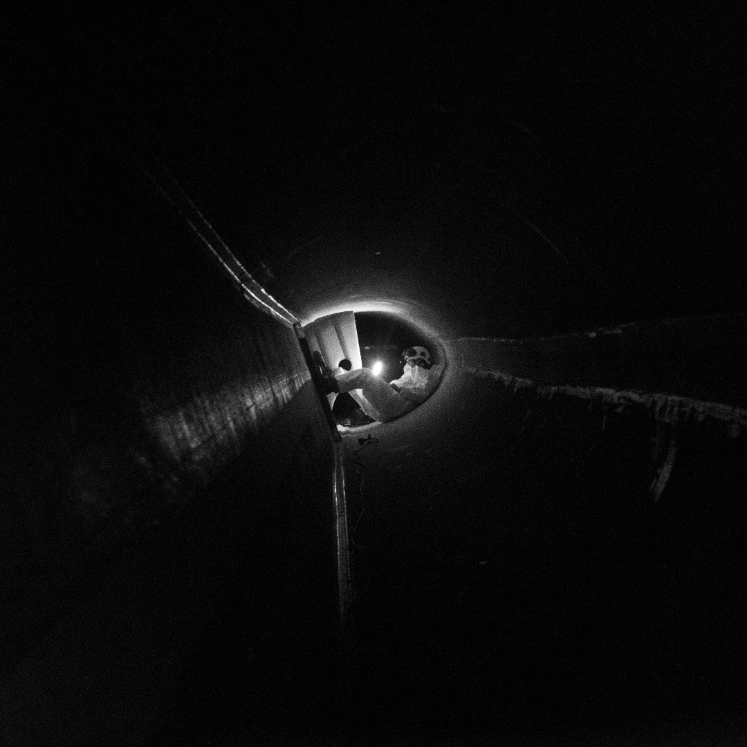 Dark tunnel interior with a lit circular opening in the distance showing a figure in protective gear.