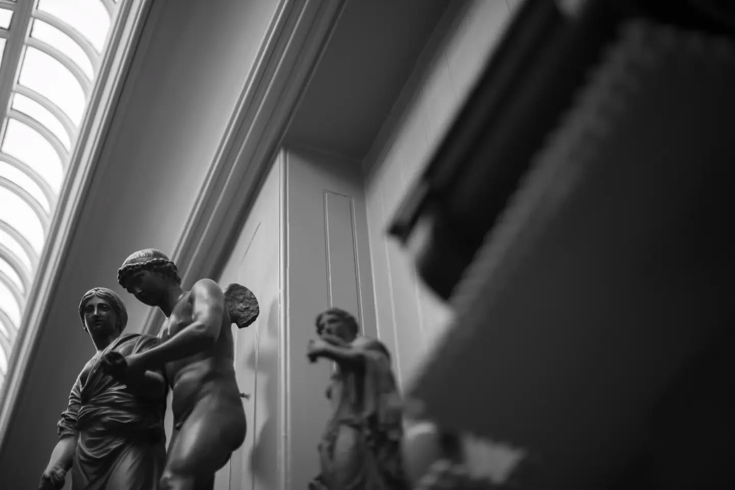 Black and white photo of classical statues in a gallery, with an arched glass ceiling visible on the left and a blurred objec
