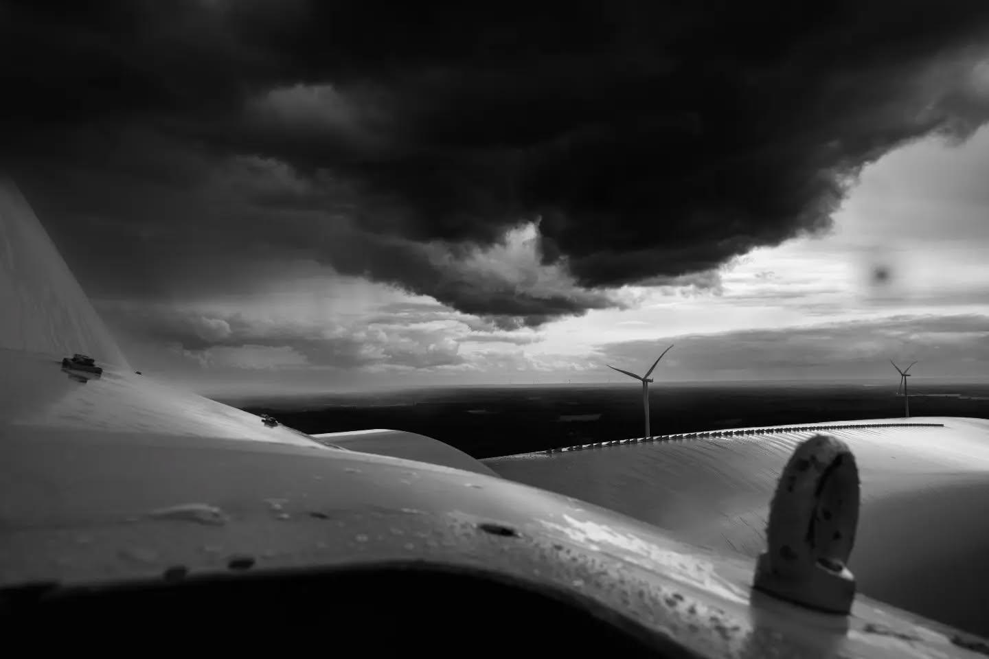 Black and white view from atop a wind turbine nacelle, with distant turbines and dark storm clouds over flat land.