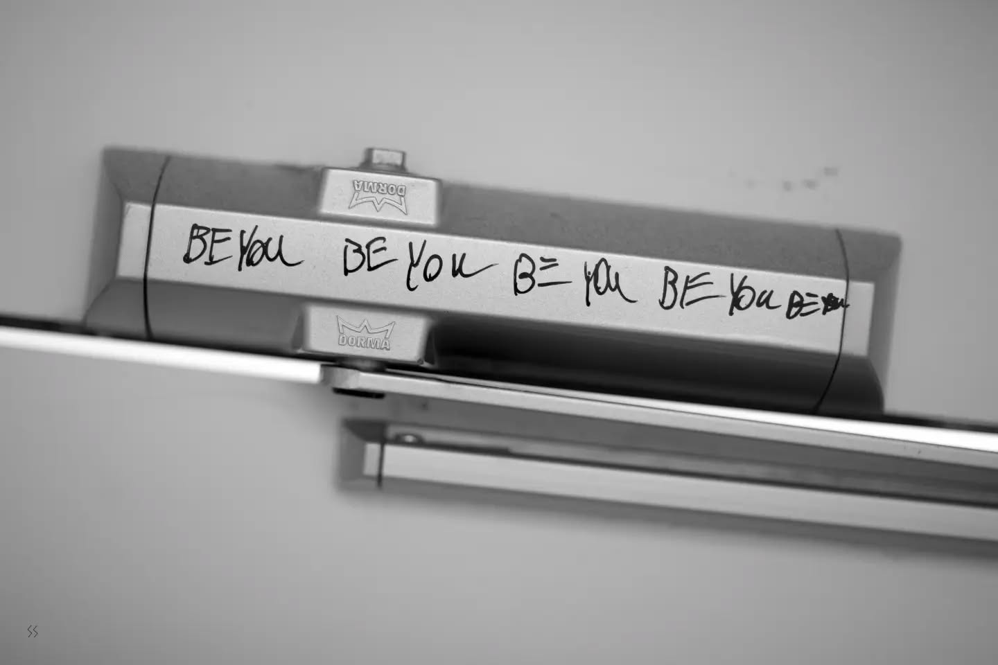 Black-and-white close-up of a Dorma door closer arm, with "BE YOU" handwritten repeatedly in marker along its metal surface.