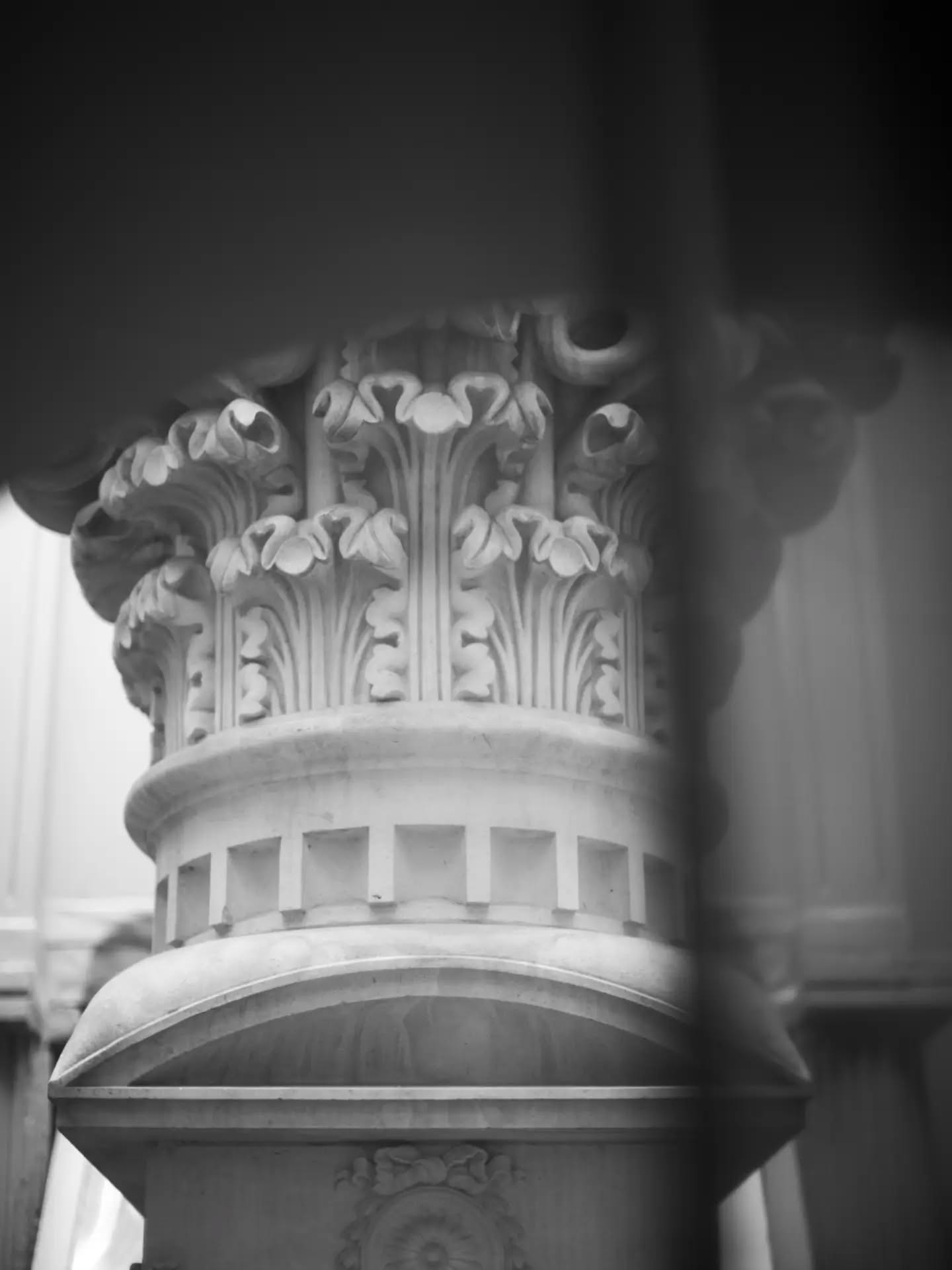 Black and white close-up of a Corinthian column capital with acanthus leaf carving, viewed through a foreground pillar edge.