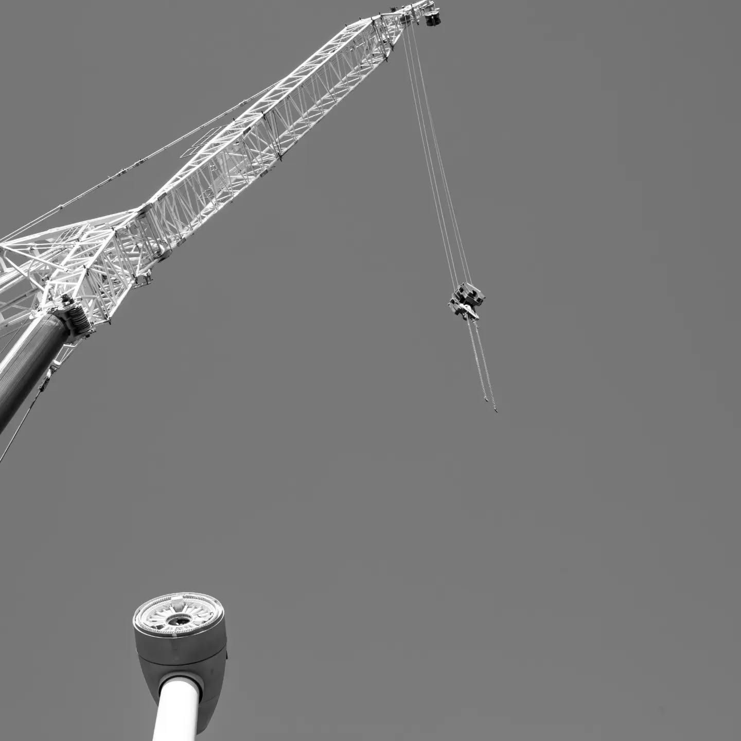 Black and white photo of a crane's lattice boom and cables against a plain sky, with a streetlamp fixture in the foreground b