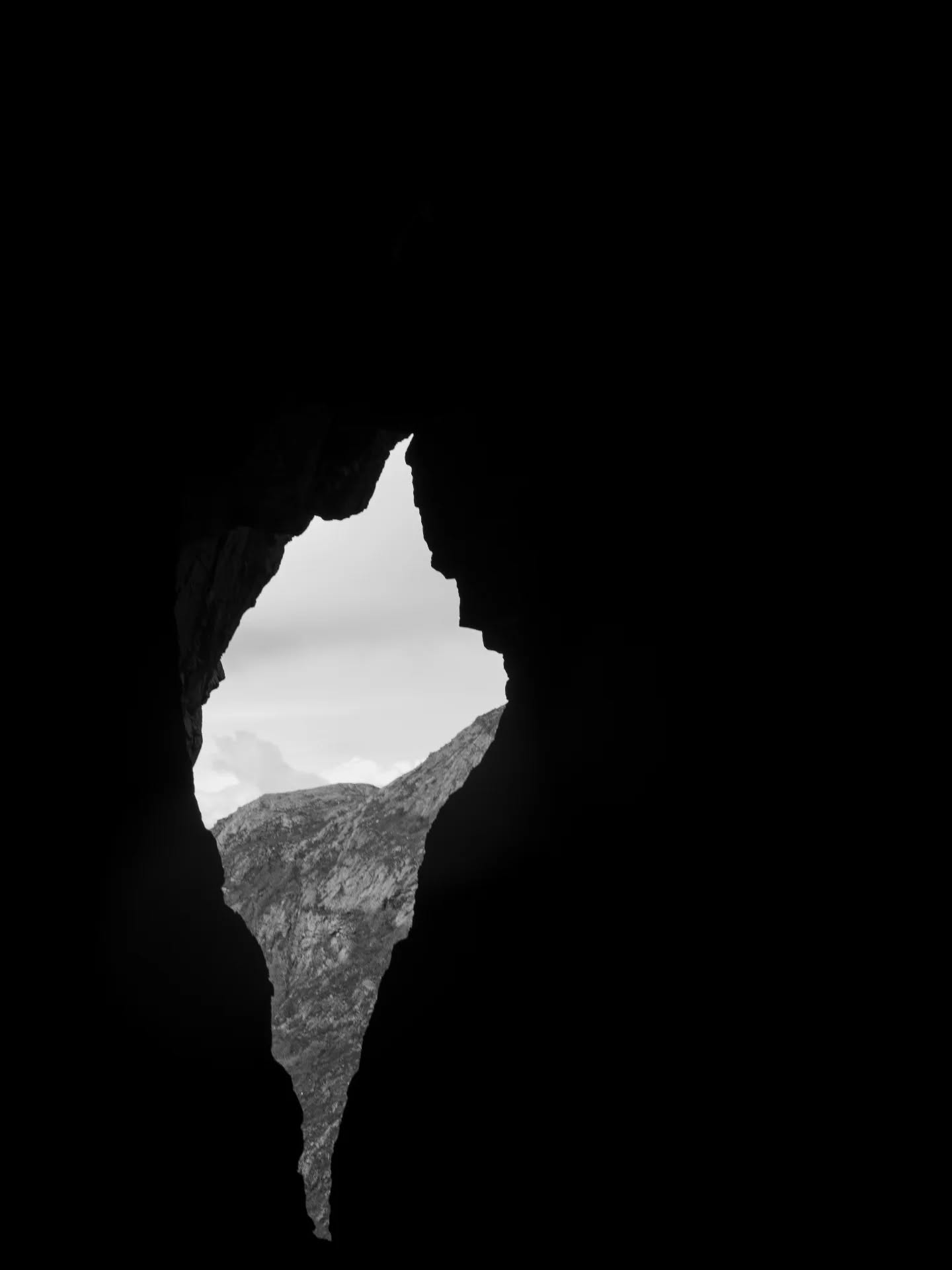 Black and white image of a jagged cave opening framed by dark rock, revealing a mountain peak and cloudy sky outside.