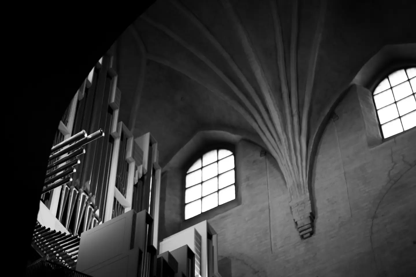 Black-and-white photo of a church interior, showing vaulted ceiling ribs, an arched window, and organ pipes at an angle.