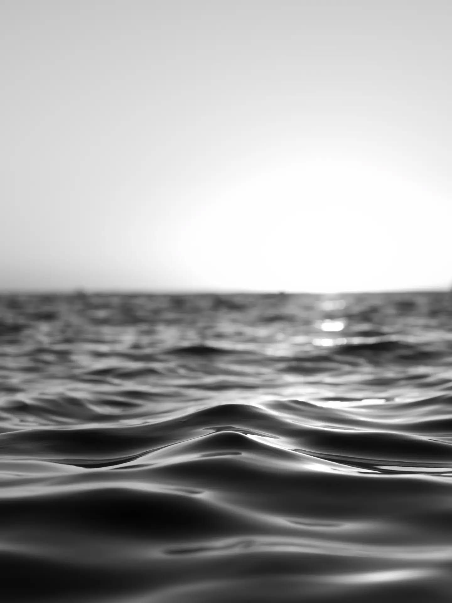 Black and white close-up of ocean water surface at eye level, with soft waves and a hazy horizon in the background.