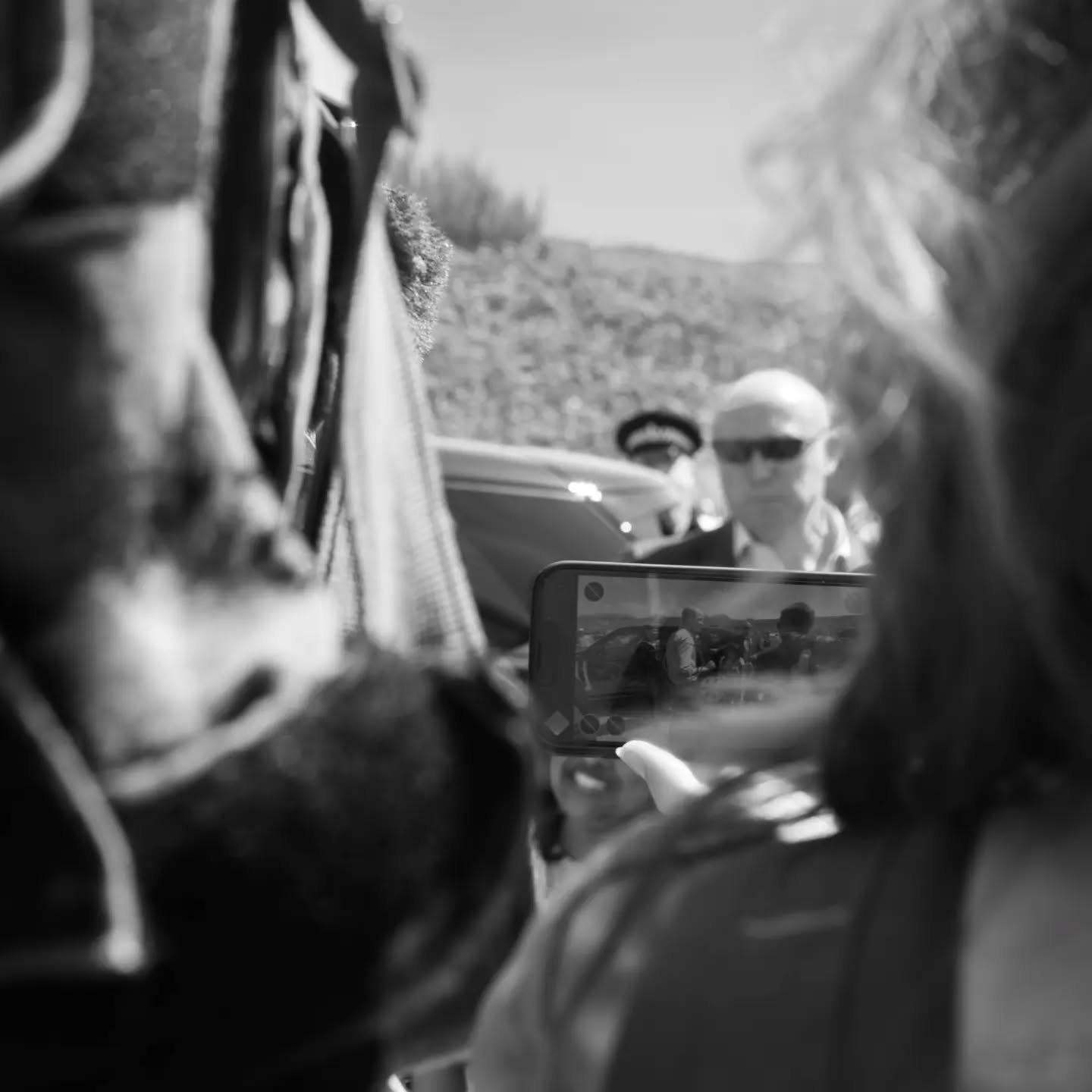 Black and white photo through a crowd of a person holding up a smartphone recording a bald man in sunglasses standing near a 