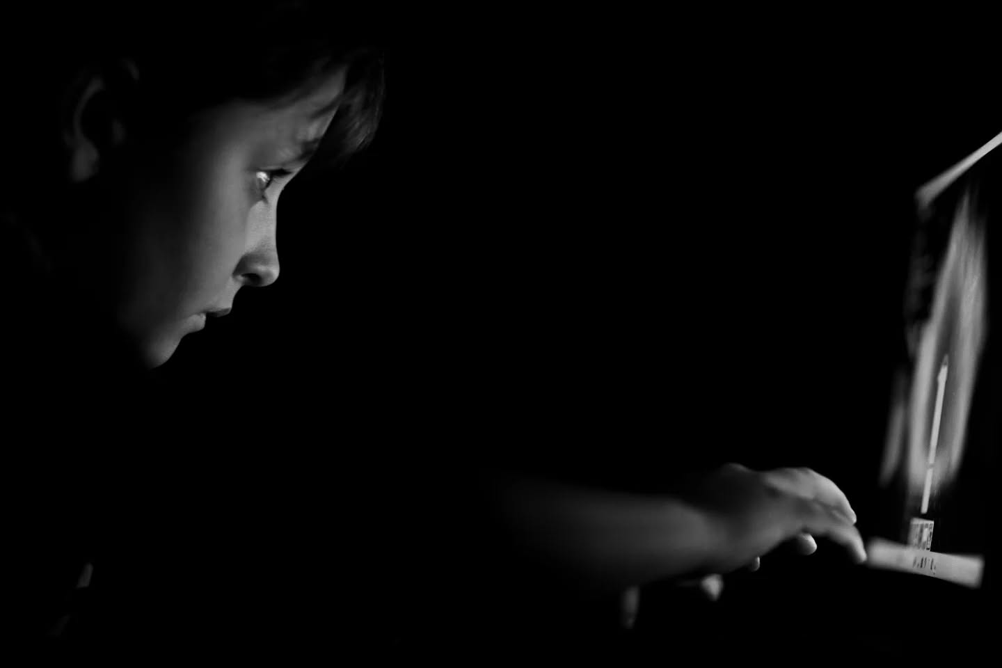 Black and white photo of a child's profile lit by a screen, hand reaching toward a laptop keyboard in darkness.