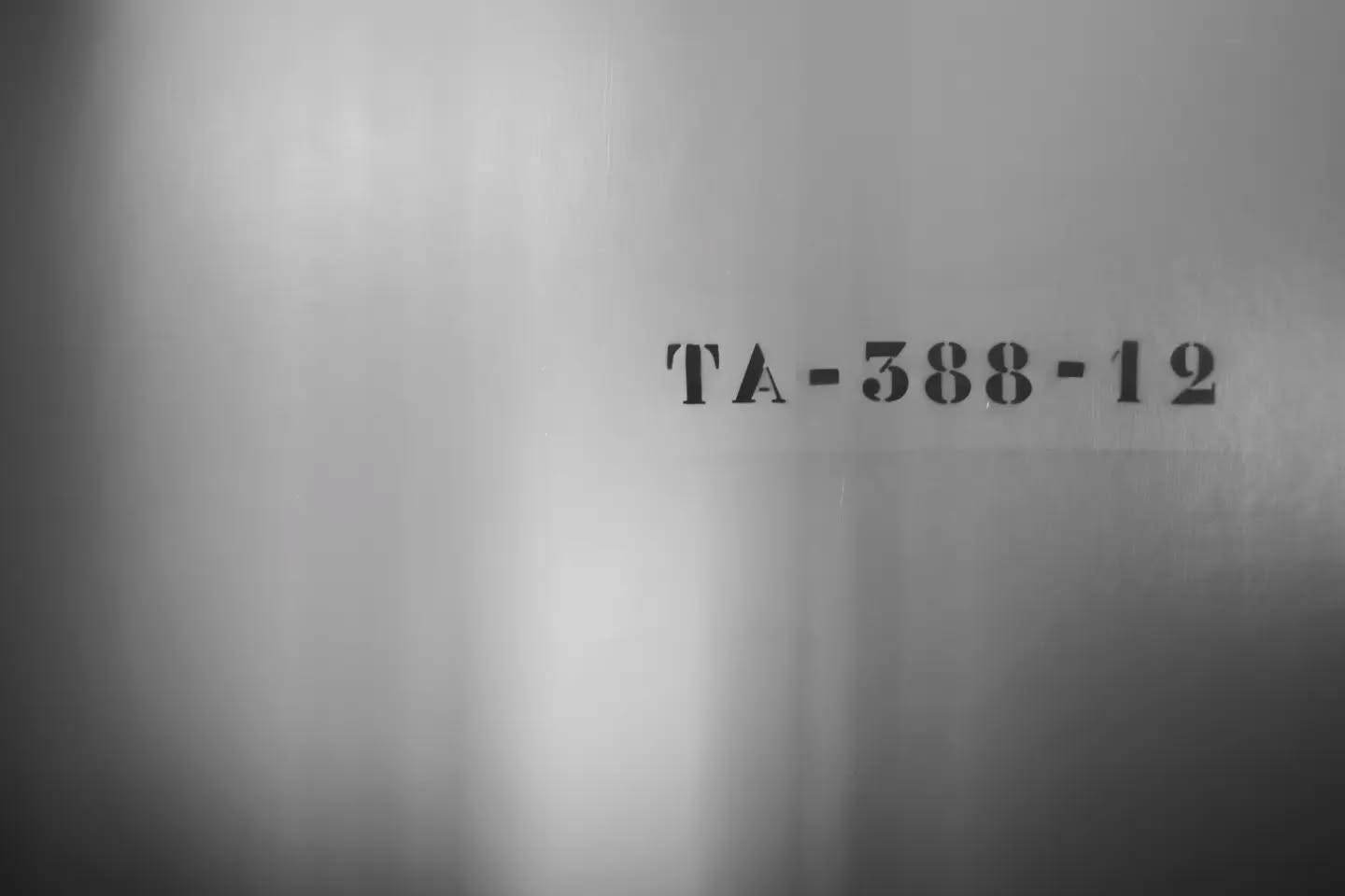 Black and white photo of a stenciled label "TA-388-12" on a smooth, softly lit metal or painted surface.