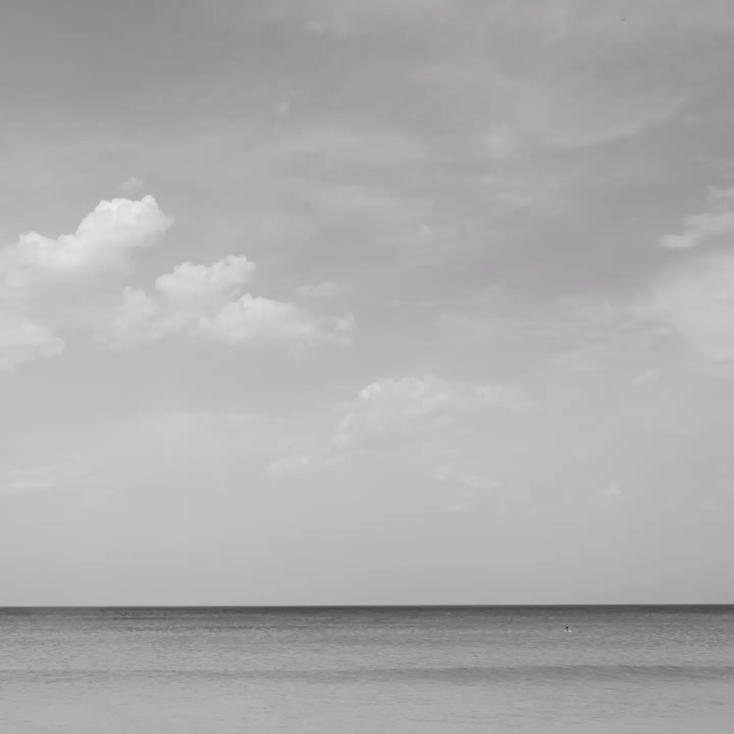Black-and-white seascape with a low horizon line, scattered clouds in a large sky, and calm water below.