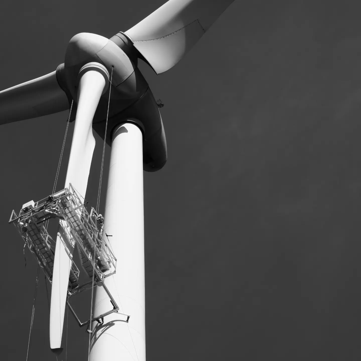 Black-and-white close-up of a wind turbine nacelle and blades with a suspended maintenance platform hanging beside the tower.