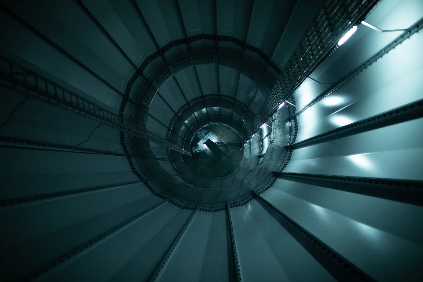 Upward view inside a spiral staircase within a cylindrical tower, metal steps and railings curving toward a lit opening above.