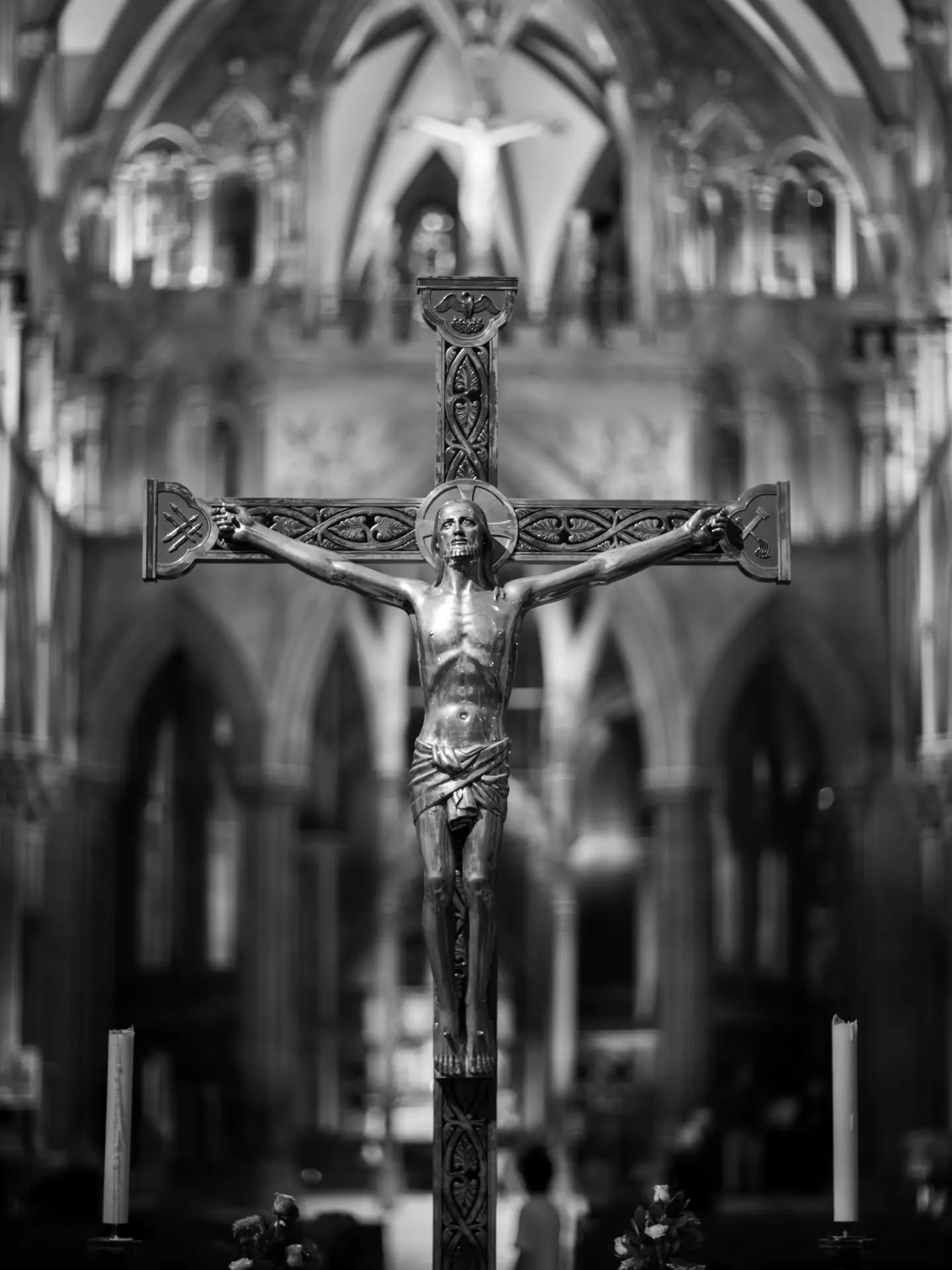 Black-and-white photo of a metal crucifix with carved cross arms, set against a blurred Gothic cathedral interior with arches and candles.