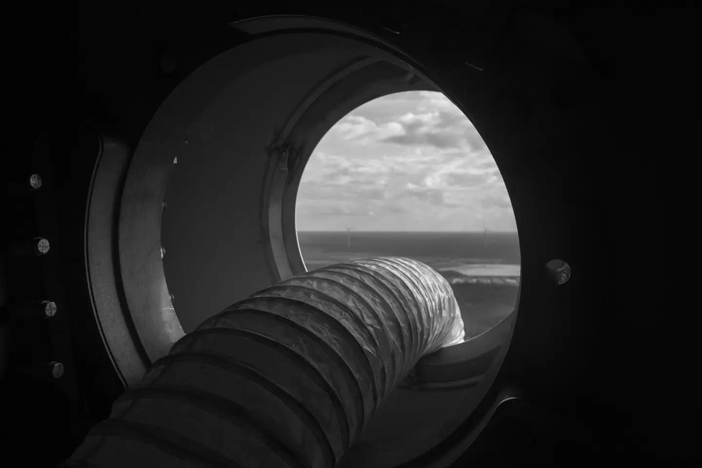 Black-and-white photo of a flexible ribbed hose extending through a round porthole window, with clouds and sea visible outside.
