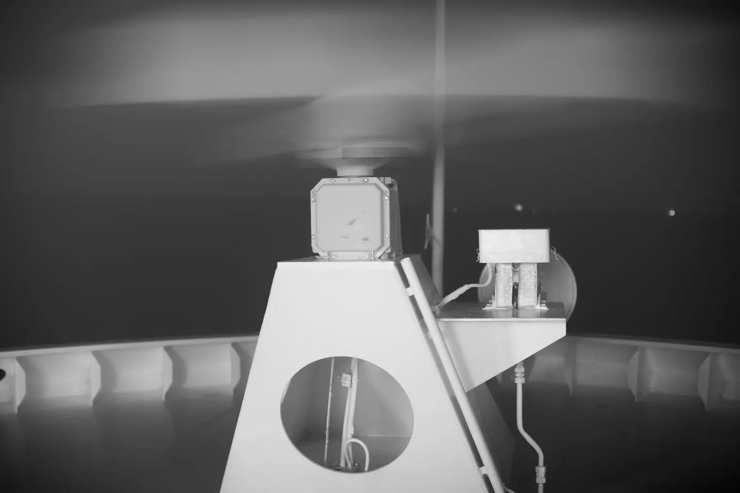Black-and-white photo of a triangular pedestal structure with a square instrument mounted on top, a smaller boxy device beside it, dark background.