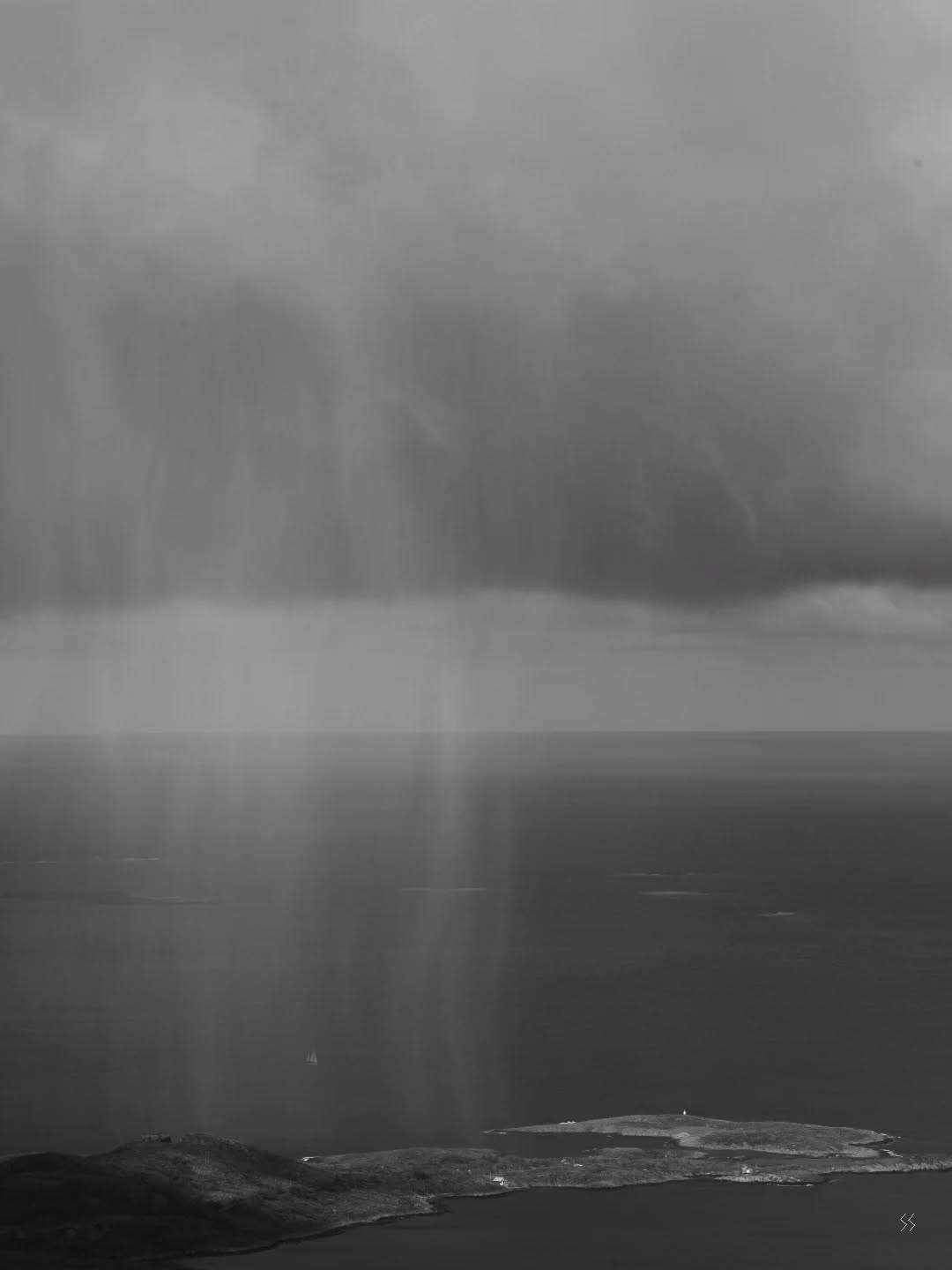 Black-and-white image of rain falling over the sea, with dark islands visible below on the horizon and a small sailboat.
