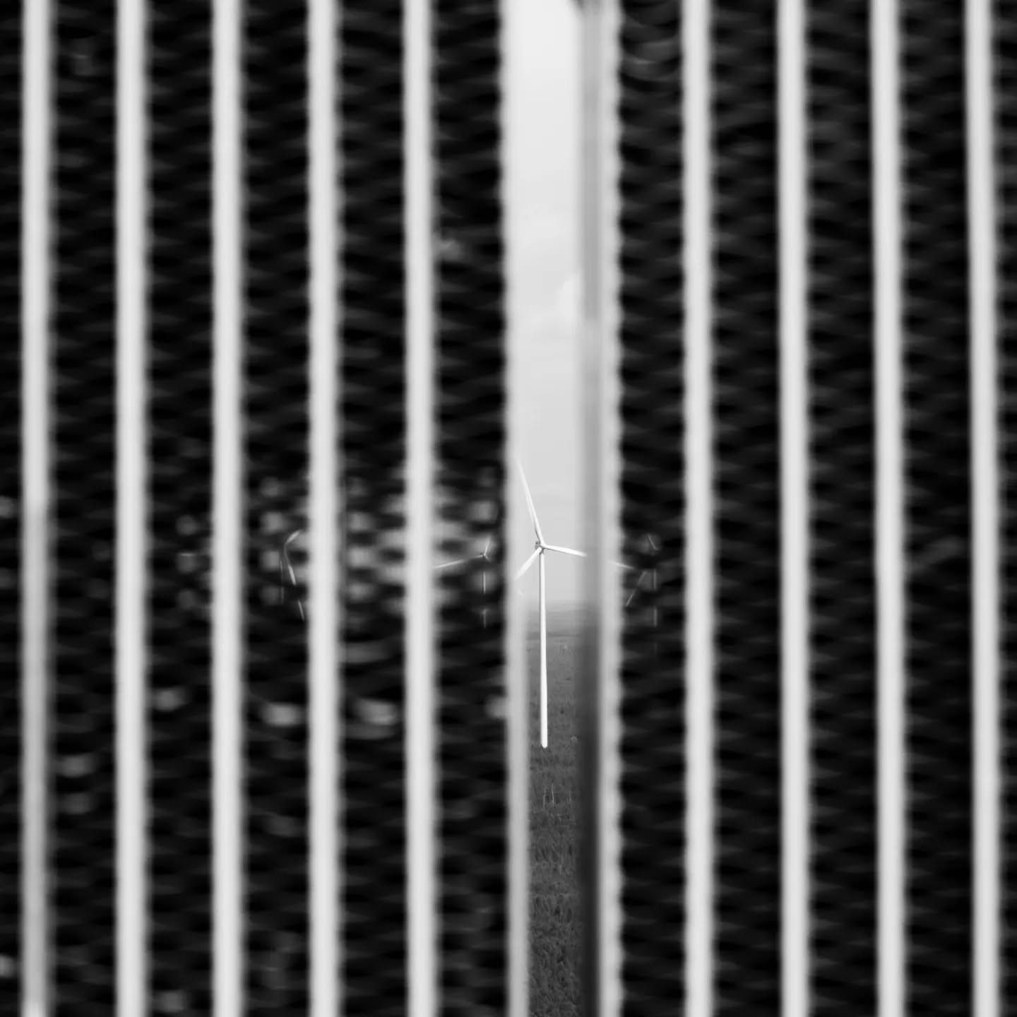 Black-and-white image of a wind turbine seen through vertical metal bars or grille slats in sharp focus, surrounding bars blu