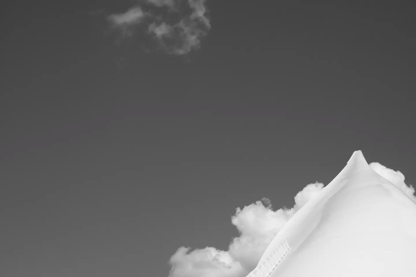 Black-and-white image of a peaked white tent roof against a gray sky with scattered clouds.