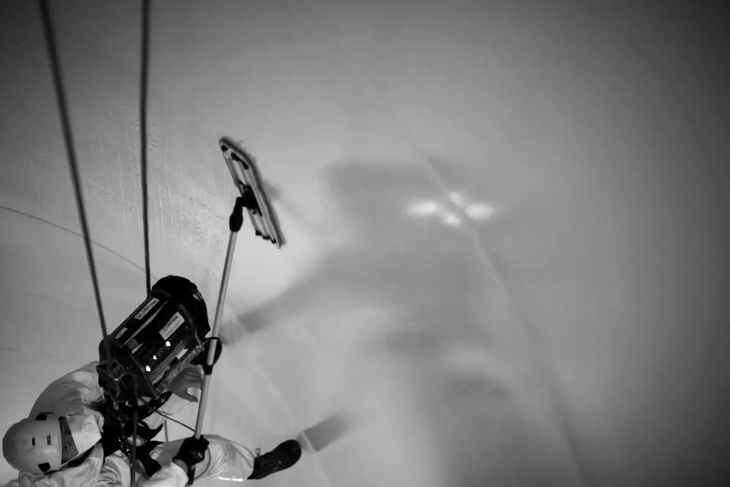 Black-and-white image of a rope-suspended worker in coveralls and helmet cleaning a curved wall with a squeegee tool.