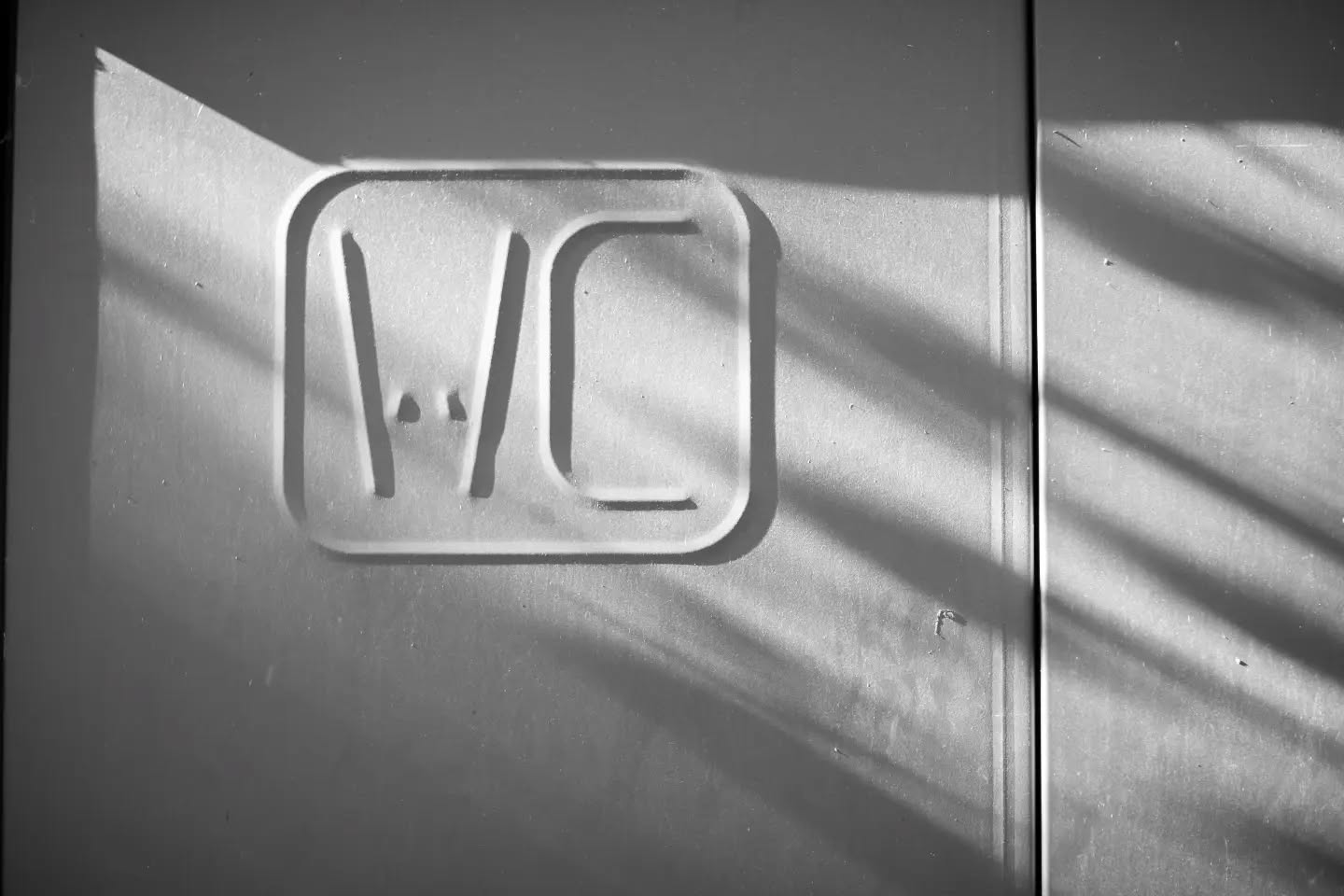Black-and-white photo of a raised "WC" sign on a textured wall, with diagonal light and shadow stripes crossing the surface.