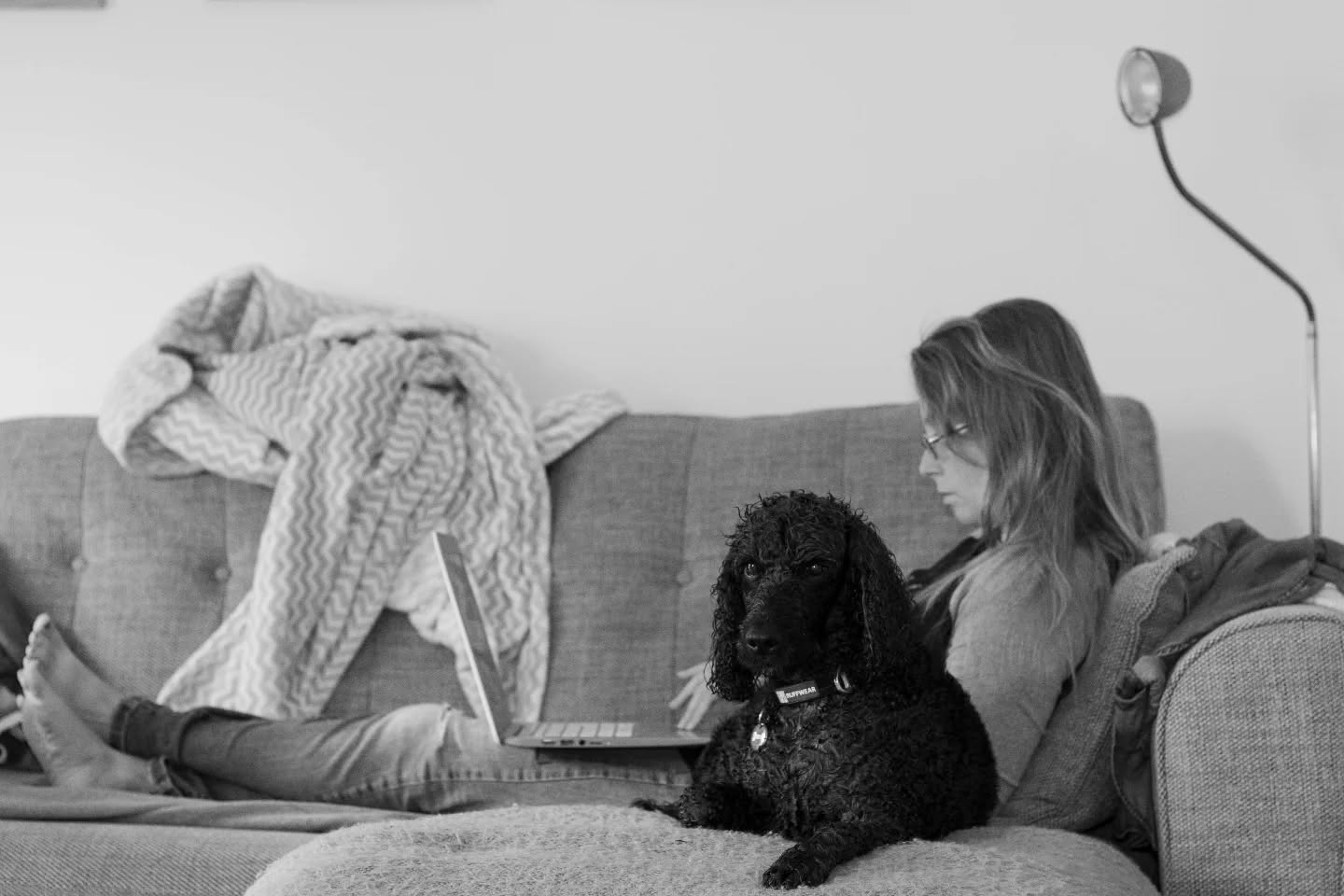 Black and white photo of a woman reclining on a couch with a laptop, a curly-haired black dog sitting beside her, legs propped up.