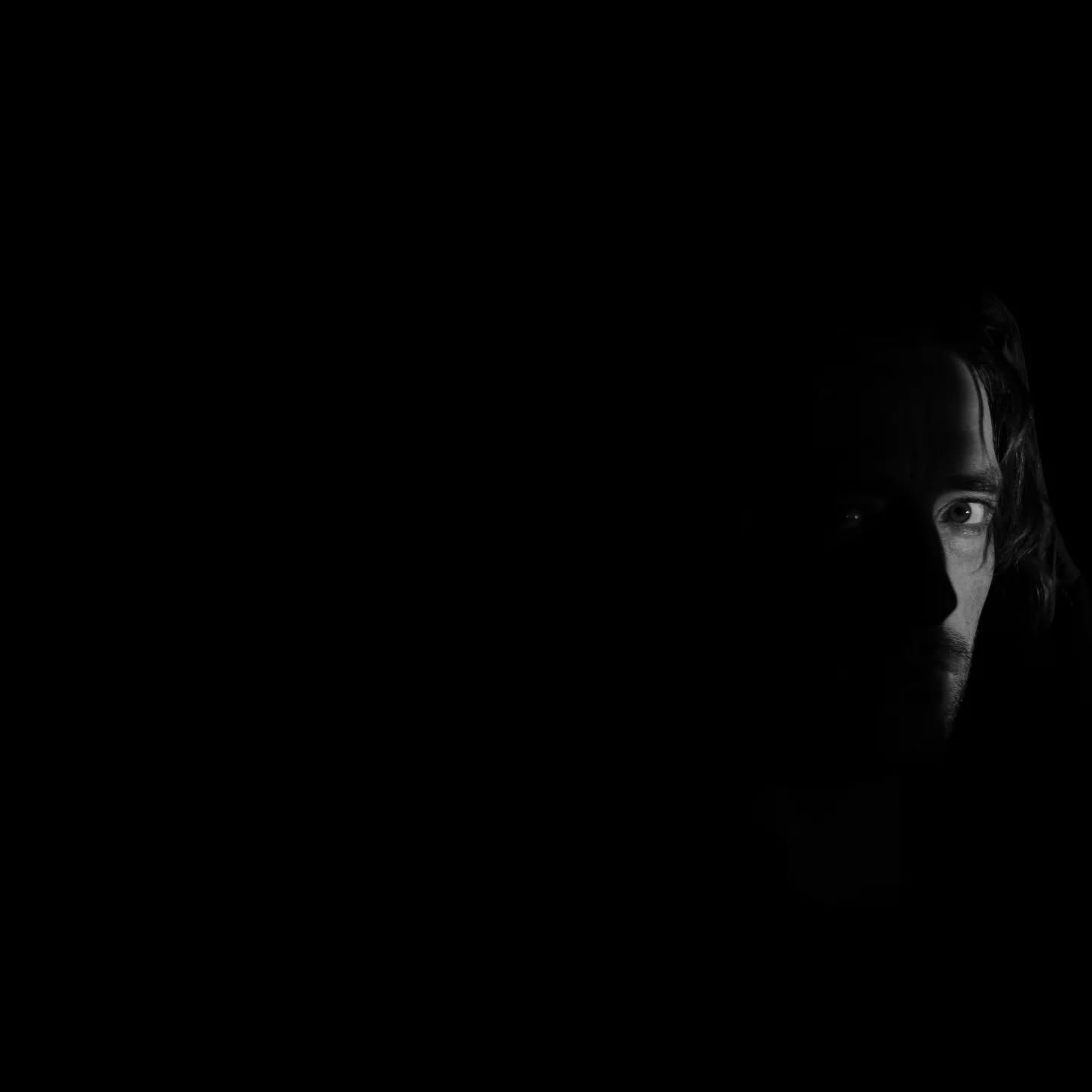 Black-and-white portrait showing the right side of a man's face partially lit, with long hair, emerging from dark shadow.