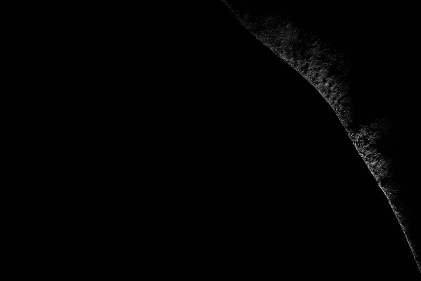 Curved illuminated edge of a textured dark sphere against black background, rest of image in deep shadow.