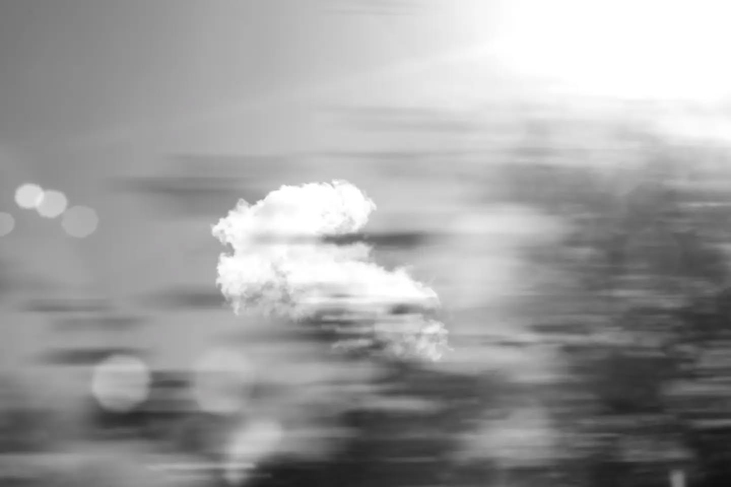 Black and white motion-blurred image of a cloud against sky, with horizontal streaks and soft circular light flares.