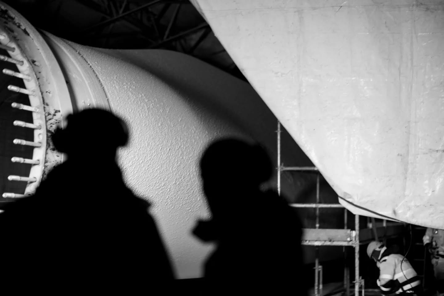 Black-and-white image showing two silhouetted figures in foreground, with large curved industrial structures and scaffolding behind them.