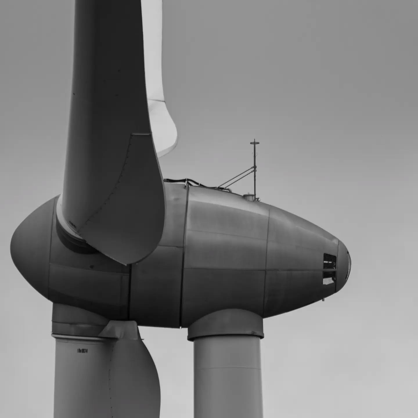 Black-and-white close-up of a wind turbine nacelle and blades against a plain gray sky.