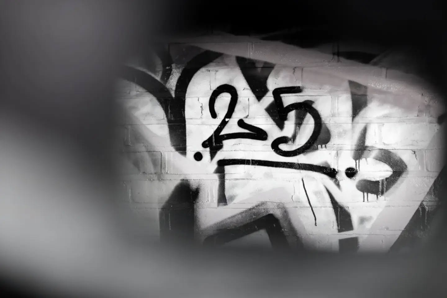 Black and white photo of graffiti tag "25" spray-painted on a brick wall, viewed through a blurred circular frame.