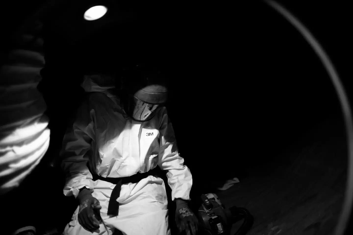 Worker in white protective suit and dark welding-style face shield inside a cylindrical structure, lit by an overhead light, black and white photo.