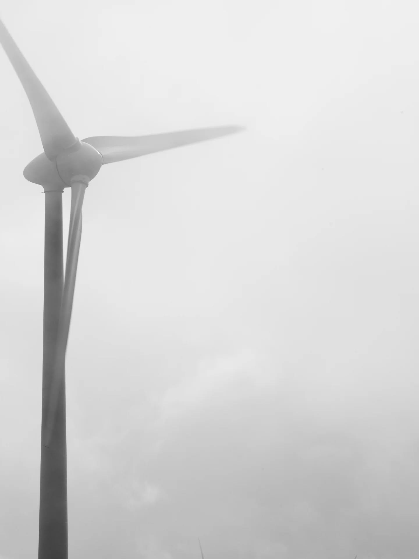 Black and white photo of a wind turbine's blades and top of tower, set against a foggy, hazy sky.