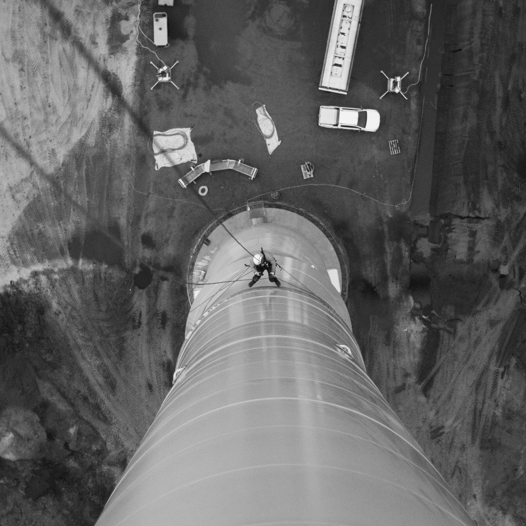 Aerial black-and-white photo of a worker rappelling on a large industrial pipe or tower, with vehicles and equipment nearby.