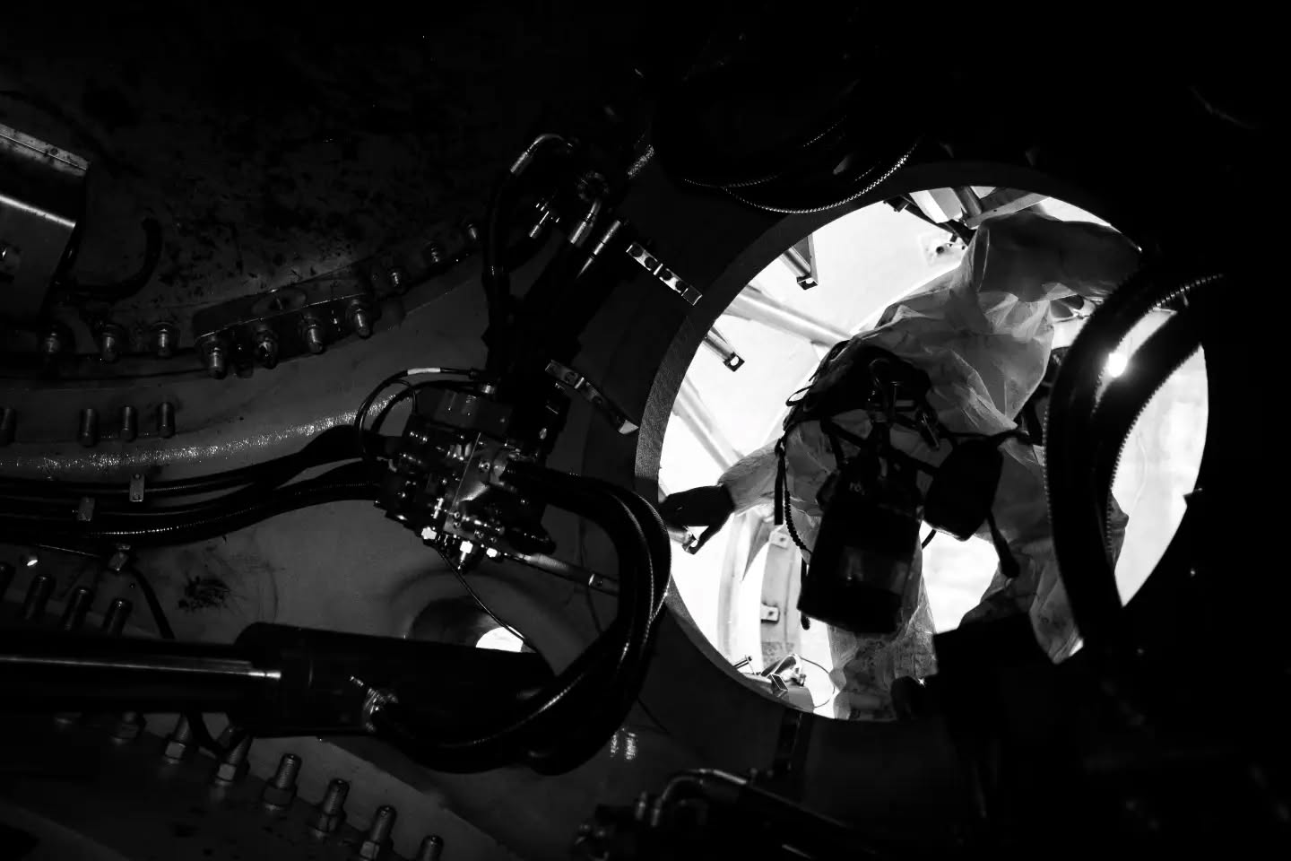 Black and white photo inside industrial machinery, showing mechanical arms and bolts, with a bright circular opening revealing a figure in protective gear.