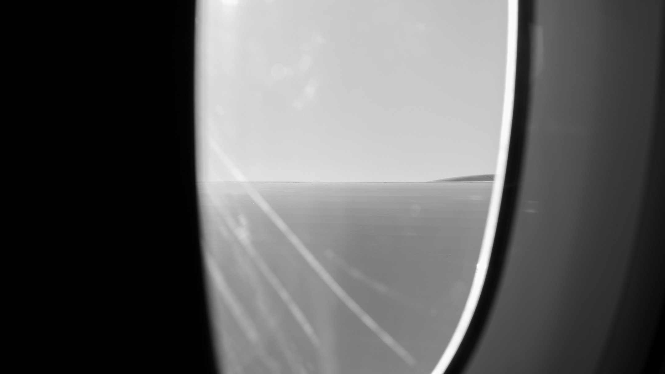 Black and white photo of a curved window edge framing a flat horizon between sky and ground, with diagonal streak marks.