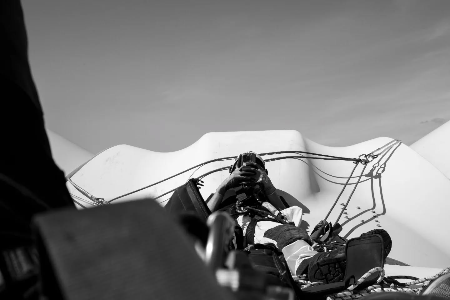 Black and white photo of a person lying back holding a camera, surrounded by paragliding harness straps and cords, with clouds behind.