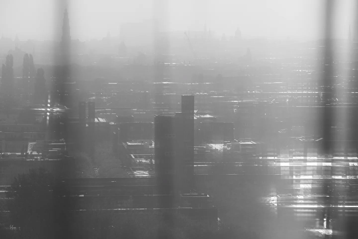 Hazy black-and-white cityscape viewed through vertical blurred foreground bars, with distant spires and rooftops fading into fog and glare.