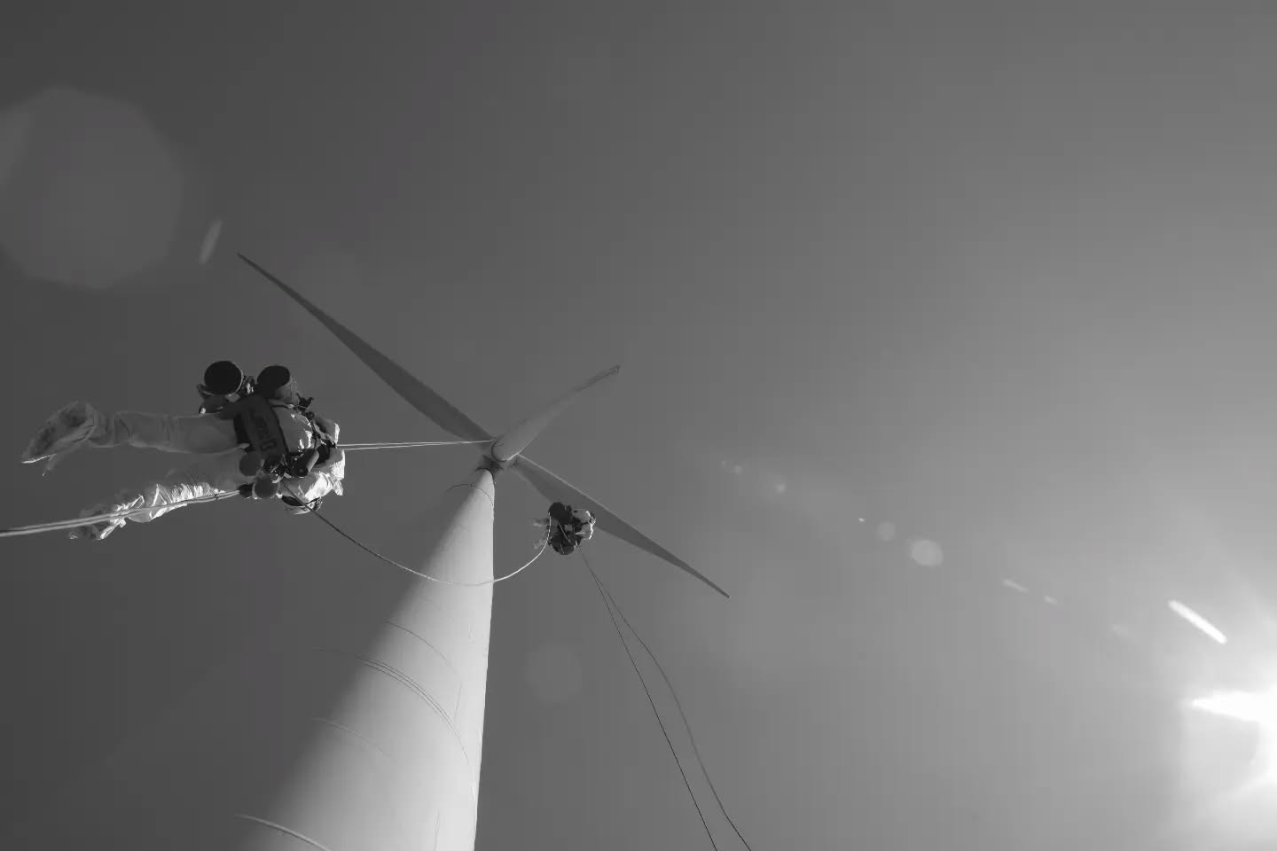 Black and white low-angle photo of a technician suspended by ropes near the hub of a wind turbine, with blades extended against a hazy sky.