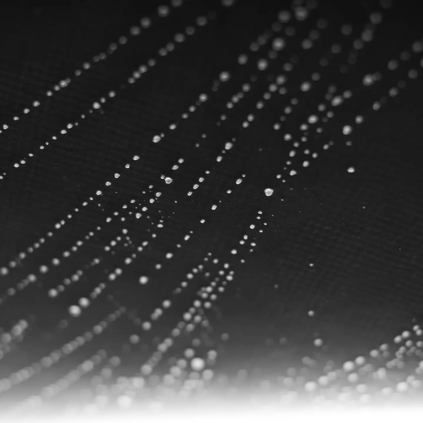 Water droplets on a dark woven fabric surface, arranged in diagonal lines, fading to white in the lower right corner.