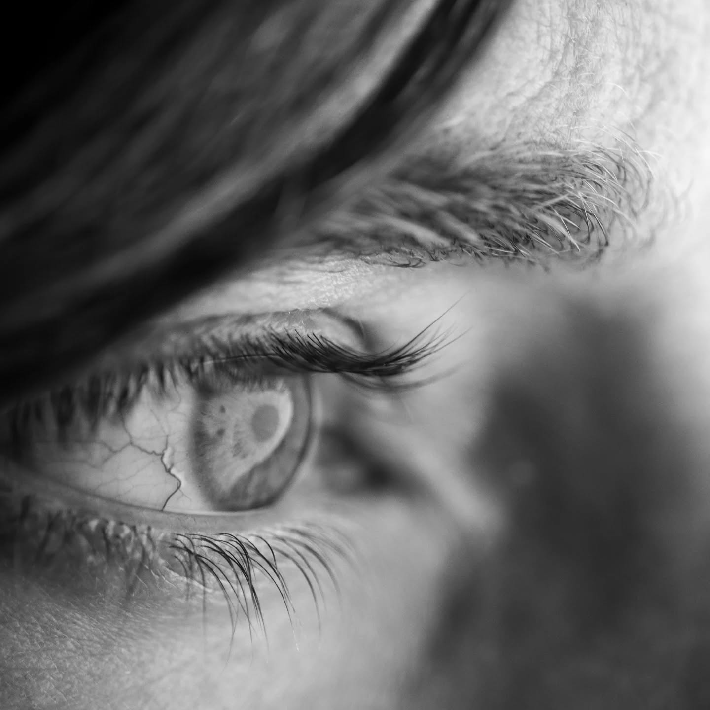 Extreme close-up black and white photo of a human eye, eyelashes, and brow, with hair falling across the forehead.