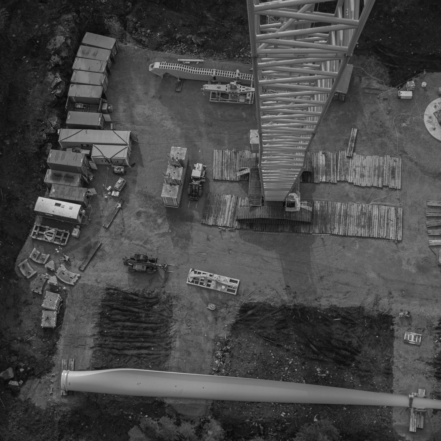 Aerial view of a wind turbine construction site with shipping containers, a wind turbine blade, crane base lattice structure, wooden mats, and construction vehicles scattered on dirt ground.