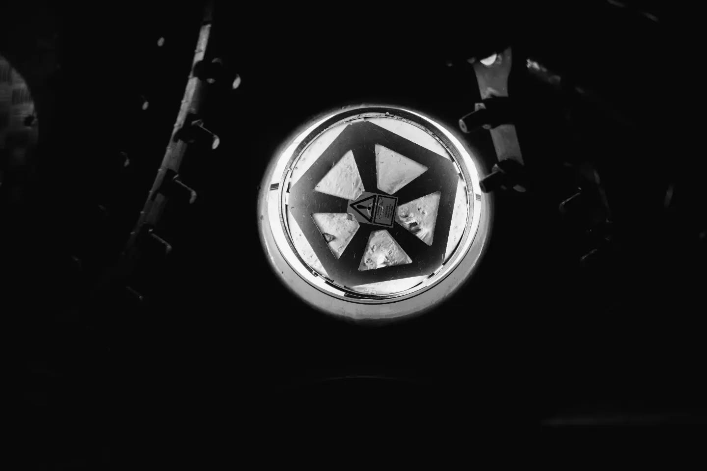 Black-and-white photo of a round hatch or vent with a triangular pinwheel pattern, illuminated and surrounded by dark machinery.